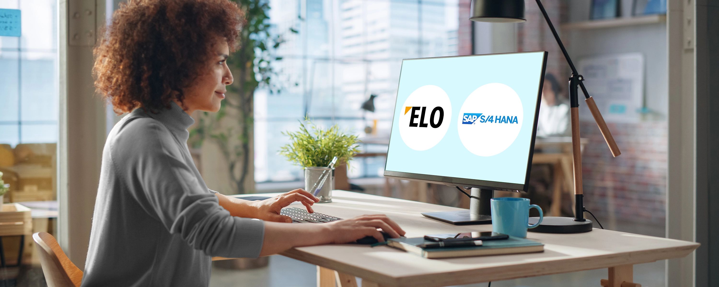 Eine Frau mit lockigen Haaren sitzt in einem modernen Büro an einem Schreibtisch und tippt. Auf dem Computerbildschirm vor ihr sind die Logos von "ELO" und "SAP S/4HANA" nebeneinander dargestellt.
