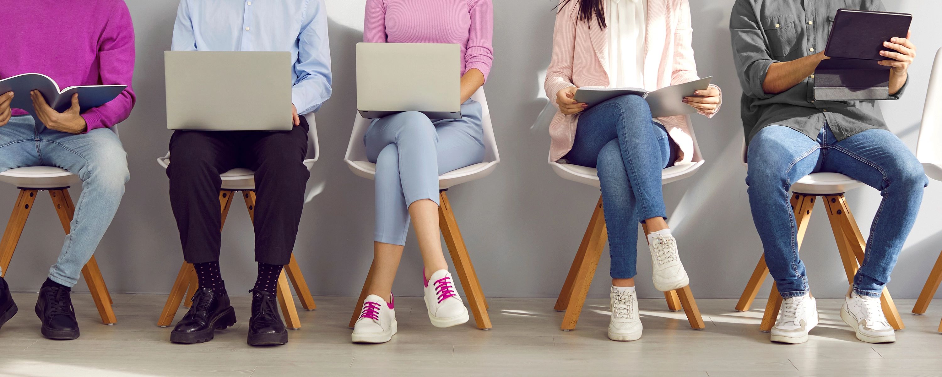 Fünf Bewerbende sitzen nebeneinander auf Stühlen mit Laptops, Tablets und Notizbüchern vor sich