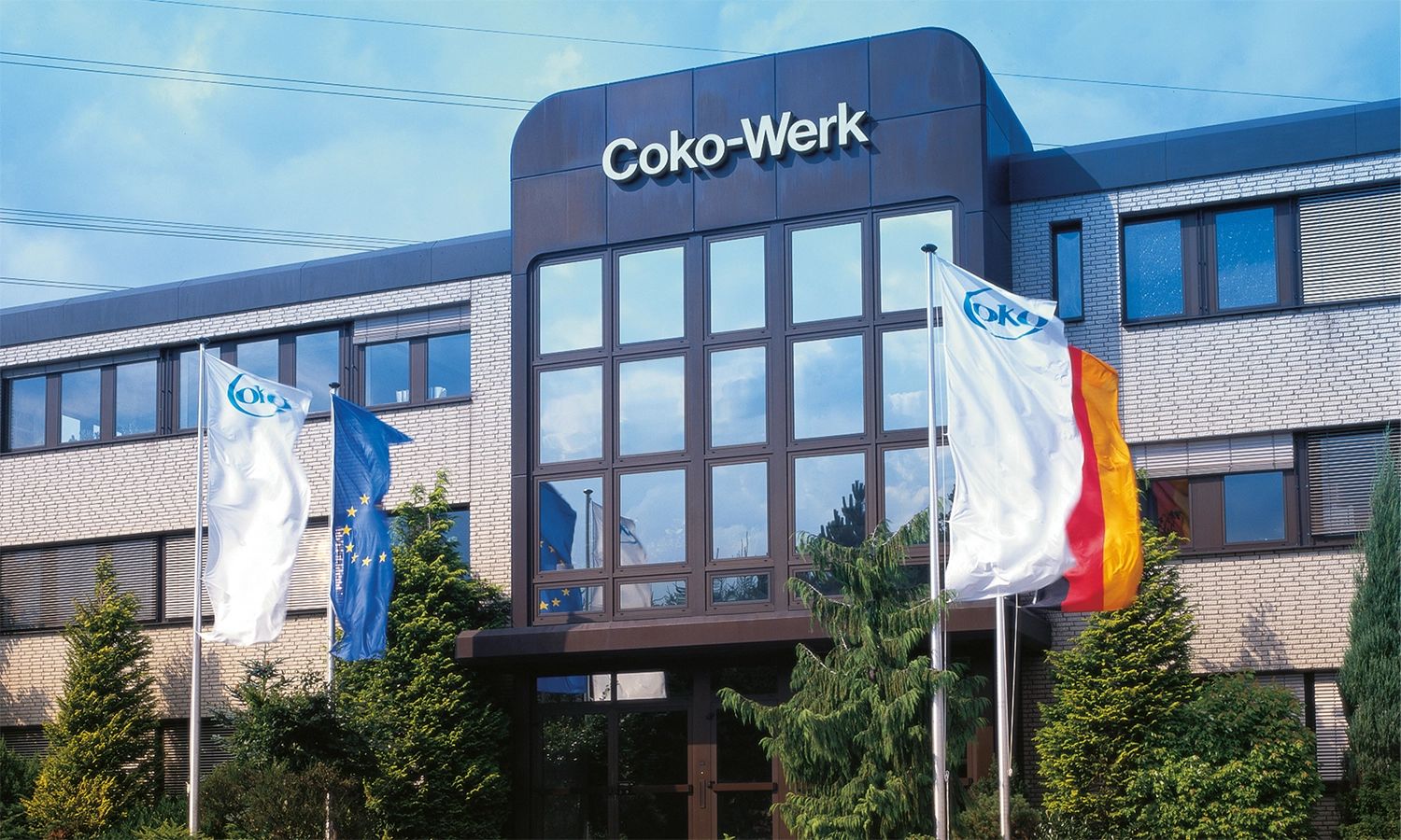 Eingangsbereich des Coko-Werks mit Unternehmens- und Länderflaggen – moderner Produktionsstandort für hochwertige Kunststofftechnik und Spritzgussteile.