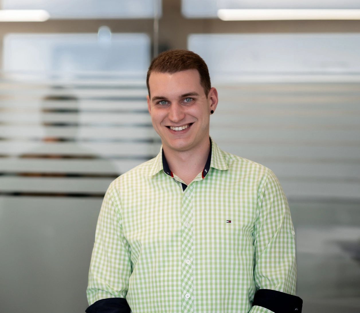 Rosenberg‑Mitarbeiter im grünen karierten Hemd vor Glaswand – Porträtfoto eines Teammitglieds des Lüftungs‑ und Klimatechnikunternehmens, das Kompetenz und Offenheit ausstrahlt.