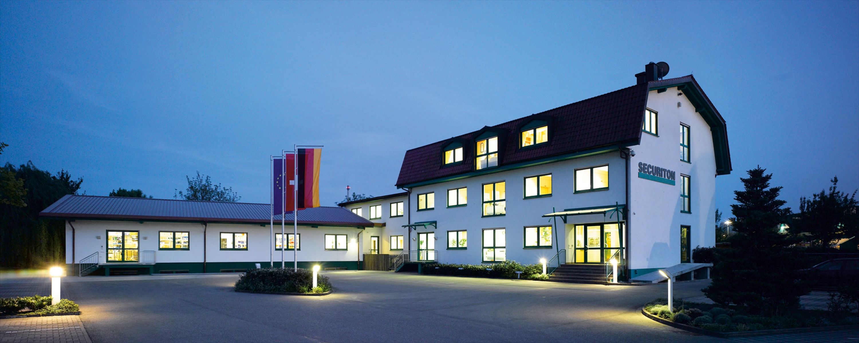 Securiton‑Firmengebäude bei Abenddämmerung – beleuchteter Hauptsitz des Sicherheits‑ und Alarmtechnikunternehmens mit Fahnenmast und modernem Eingangsbereich.