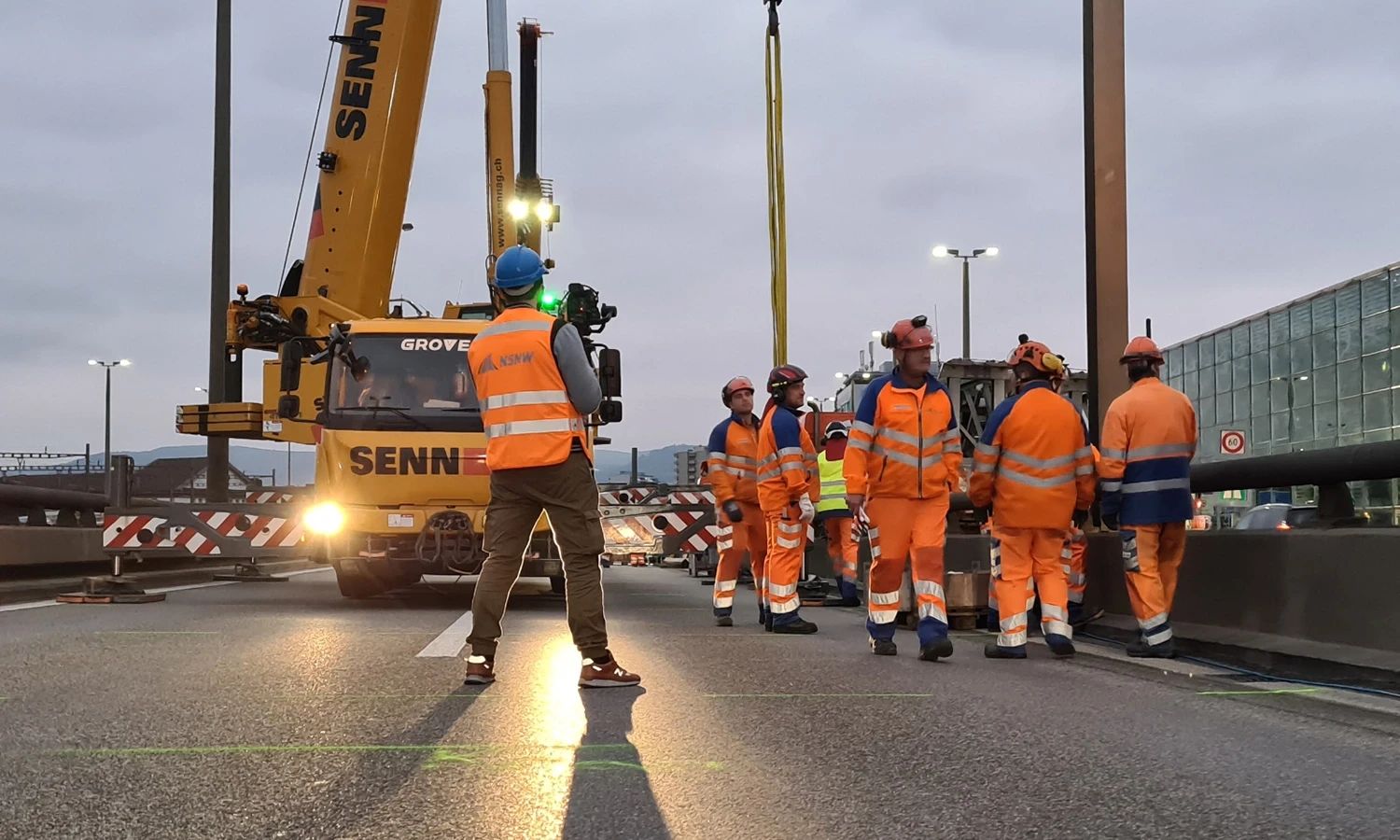 NSNW‑Bauteam bei Brückenarbeiten – Straßen‑ und Tiefbauarbeiter in Sicherheitskleidung koordinieren Hebeeinsatz mit Kran auf Baustelle.