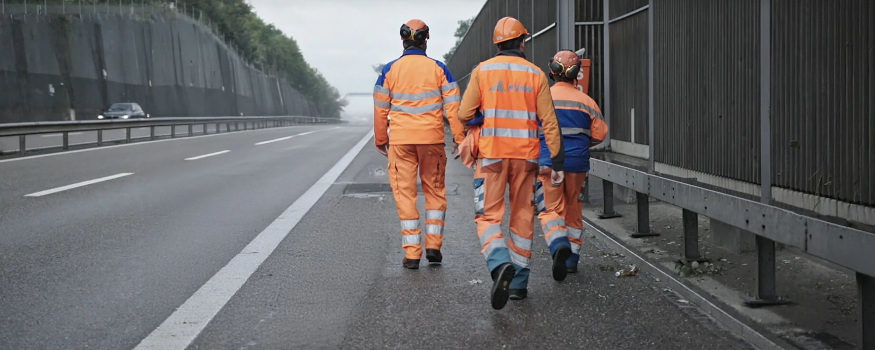 NSNW‑Mitarbeiter in Warnkleidung gehen entlang einer Autobahn – Straßenbau‑ und Instandhaltungsteam bei Arbeiten an Lärmschutzwand.