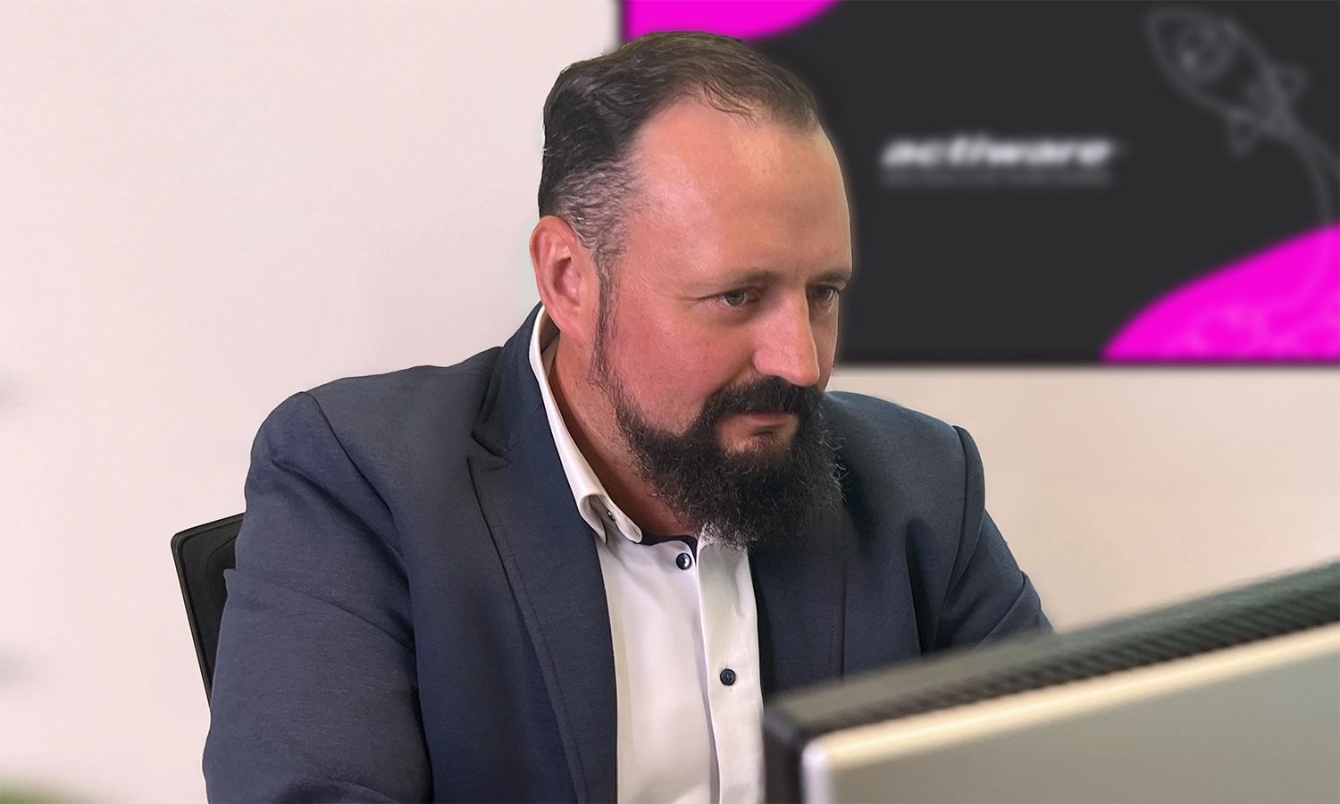 Ein Mitarbeiter schaut konzentriert auf seinen Monitor, im Hintergrund hängt ein weiterer Bildschirm mit dem ACTIWARE-Desktop-Hintergrund.