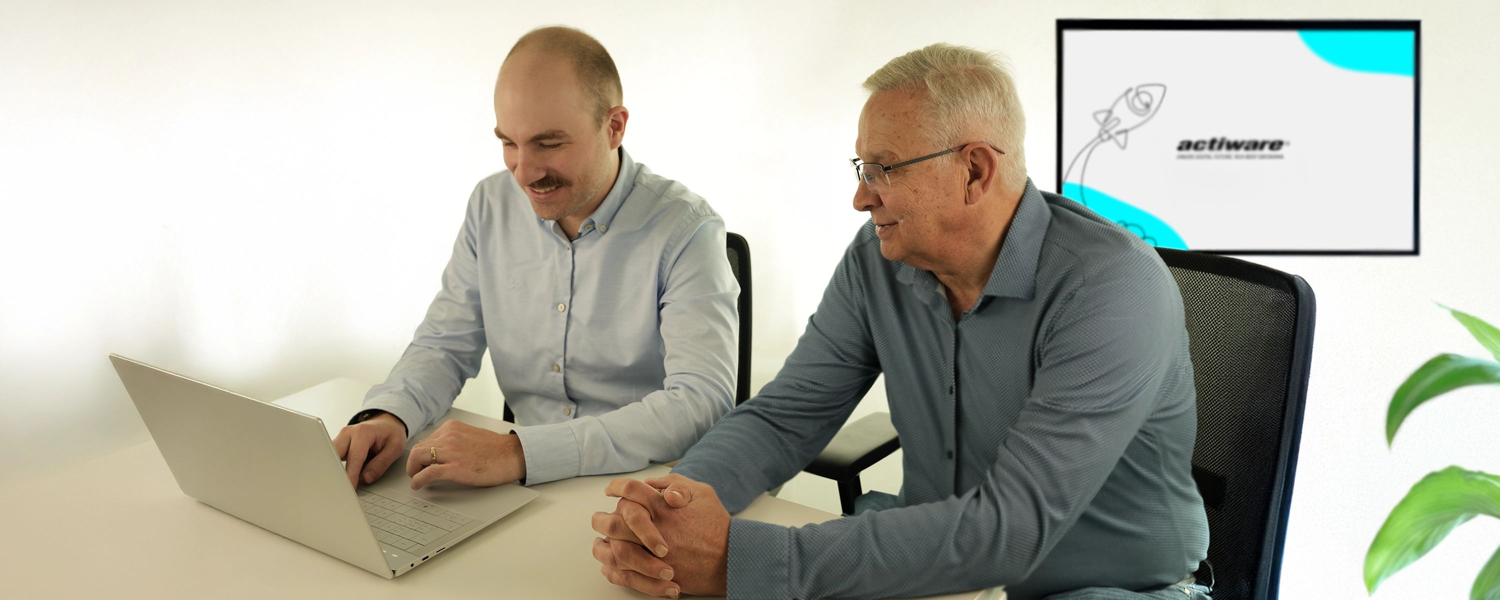 Zwei Männer sitzen gemeinsam an einem Schreibtisch und schauen auf einen Laptop. Beide lächeln und wirken konzentriert. Im Hintergrund ist ein Bildschirm mit dem Logo „actiware“ zu sehen.