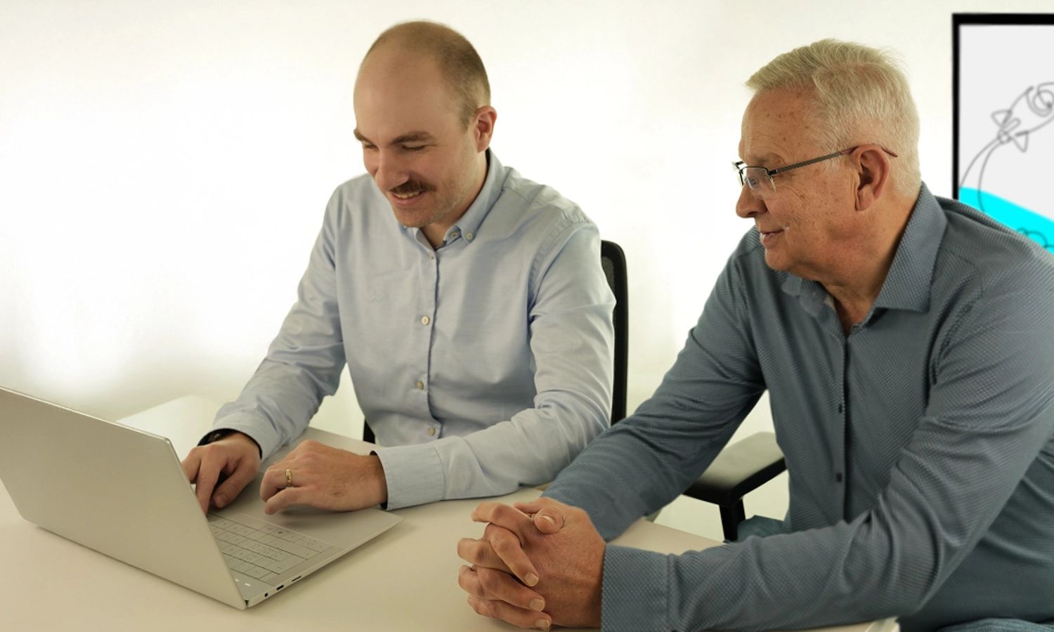 Zwei Männer sitzen gemeinsam an einem Schreibtisch und schauen auf einen Laptop. Beide lächeln und wirken konzentriert. Im Hintergrund ist ein Bildschirm mit dem Logo „actiware“ zu sehen.